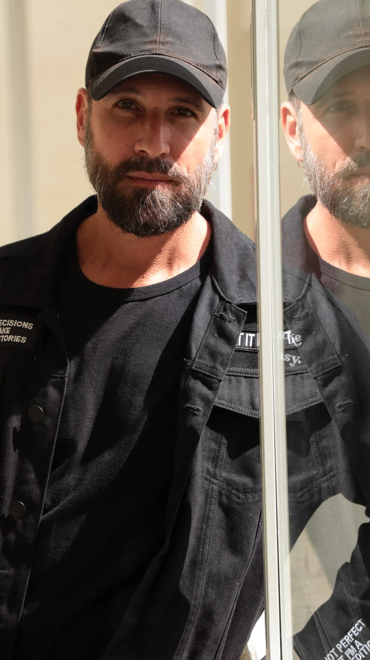 Vini Cavalieri de boné e jaqueta jeans escura, ao lado de um espelho