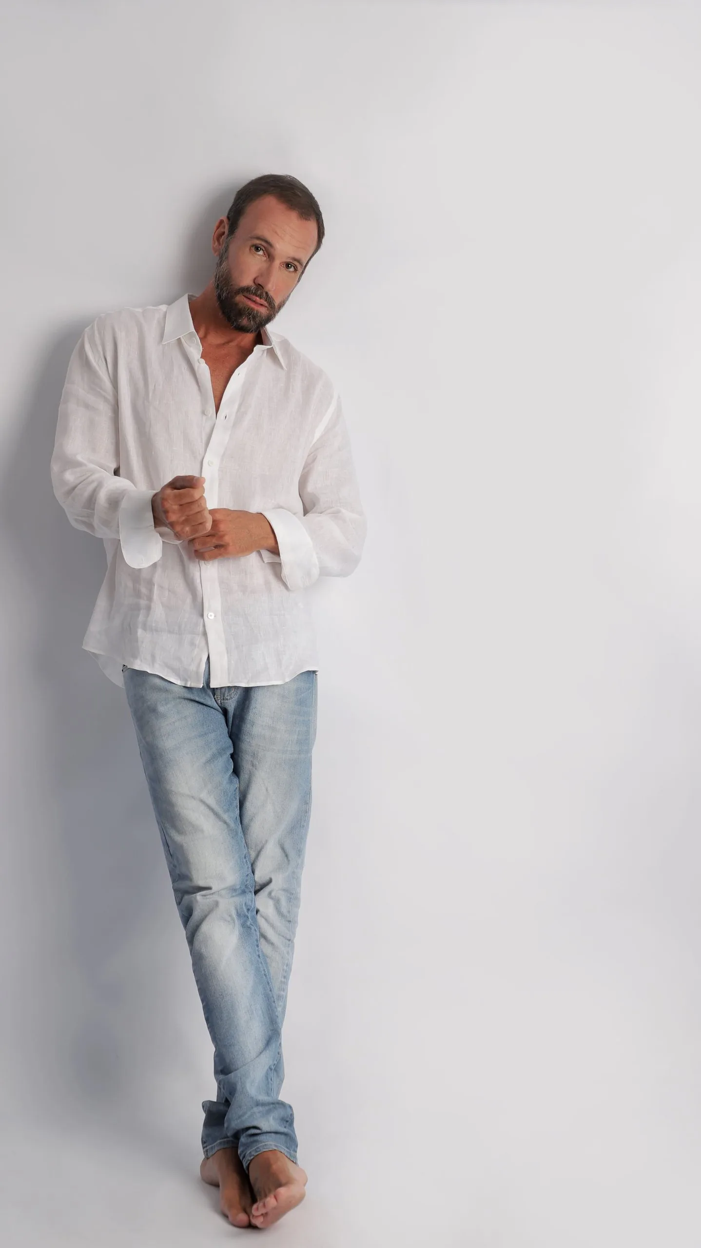 Vini Cavalieri de corpo inteiro, de camisa branca de linho e jeans, encostado a uma parede clara