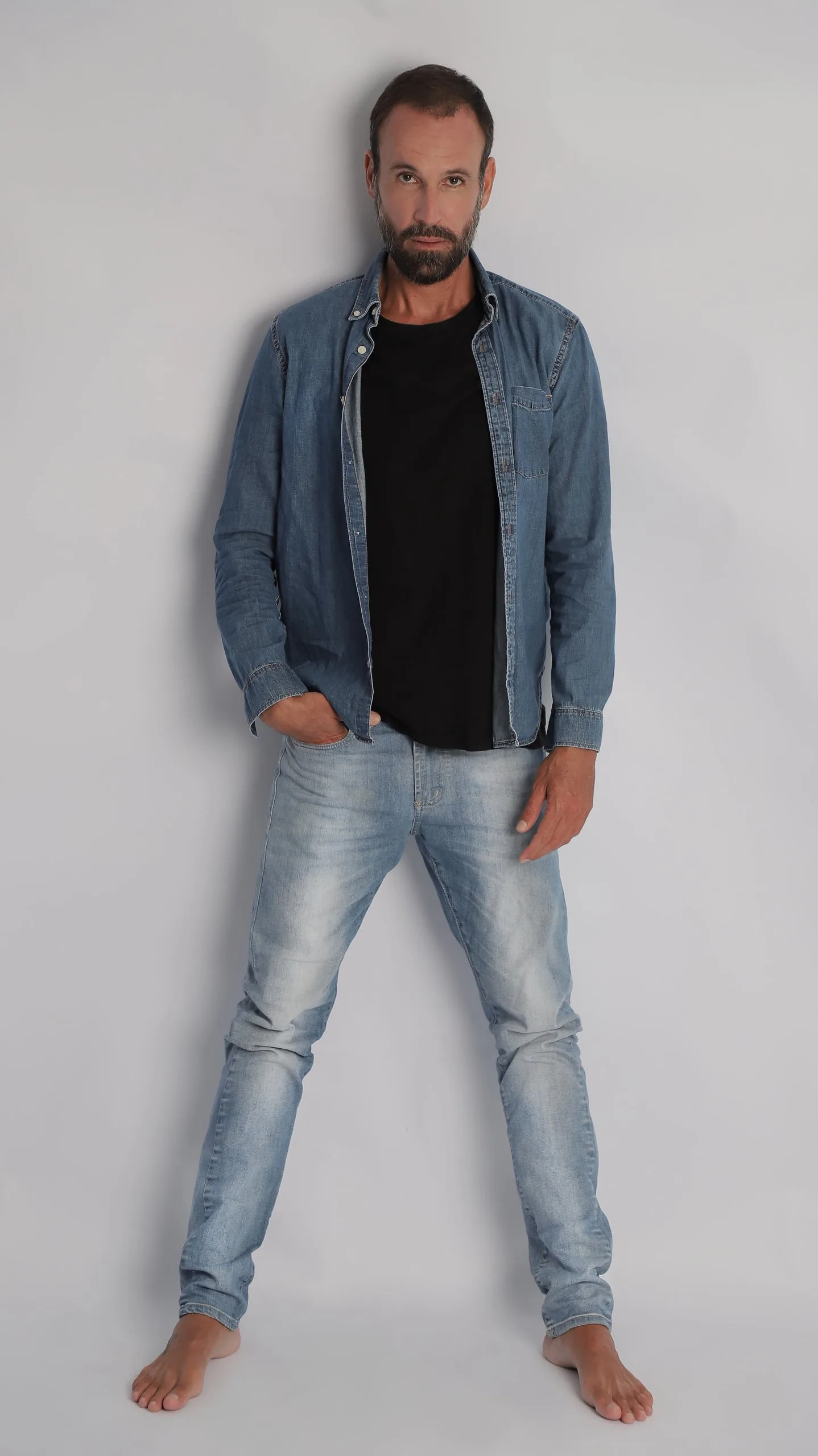 Vini Cavalieri de corpo inteiro, de camisa jeans e pés descalços, em fundo claro