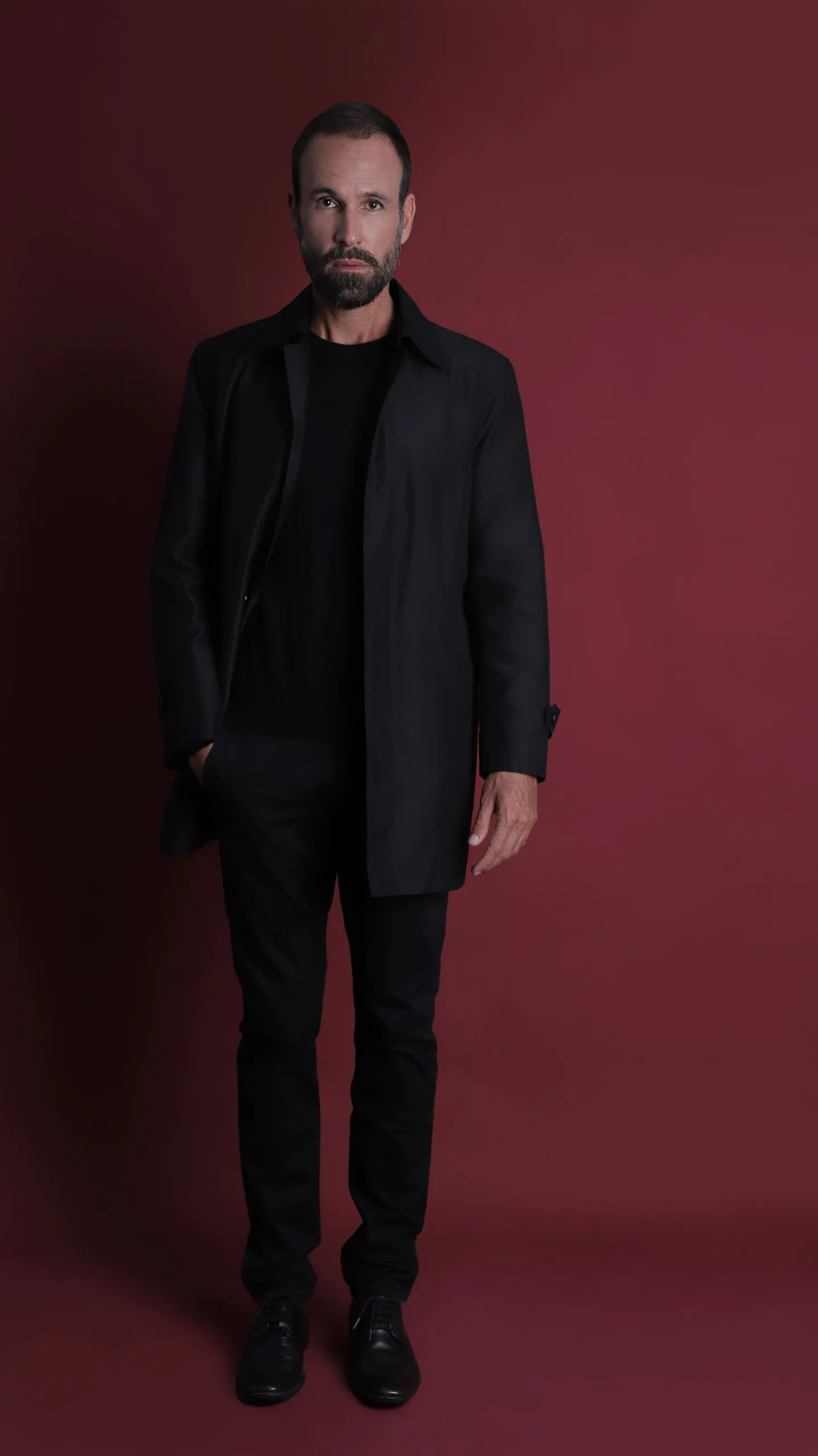 Vini Cavalieri de corpo inteiro, todo de preto, em fundo vinho