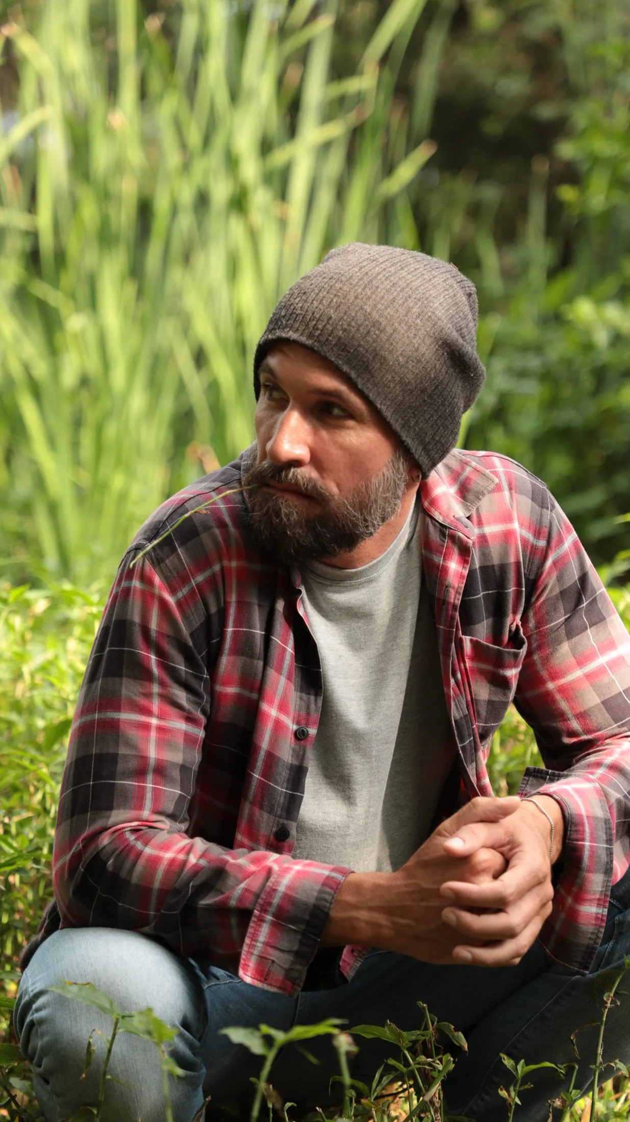 Vini Cavalieri de gorro e camisa xadrez, agachado entre a vegetação