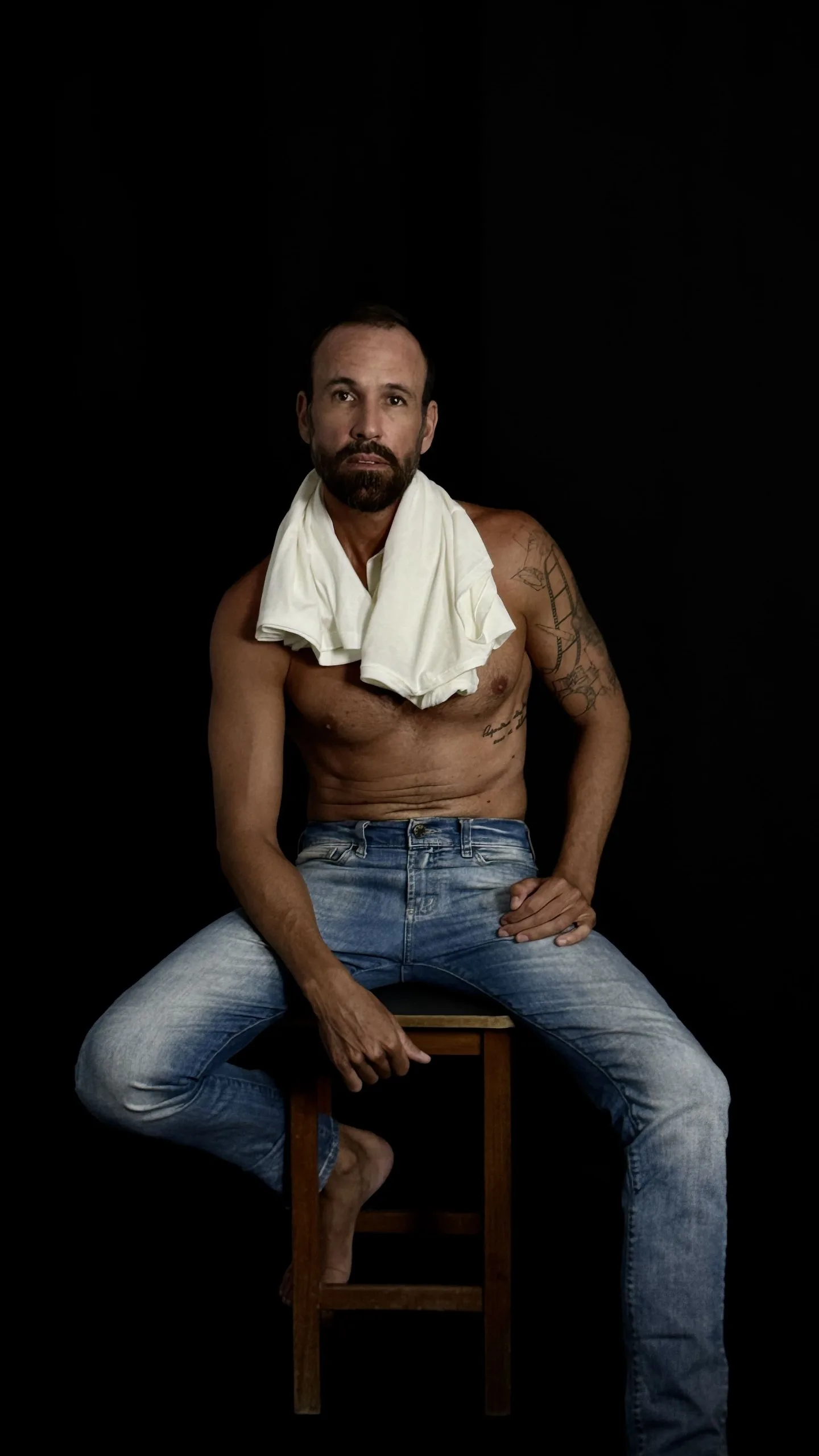 Vini Cavalieri sem camisa, com uma toalha no pescoço, sentado num banco em fundo preto