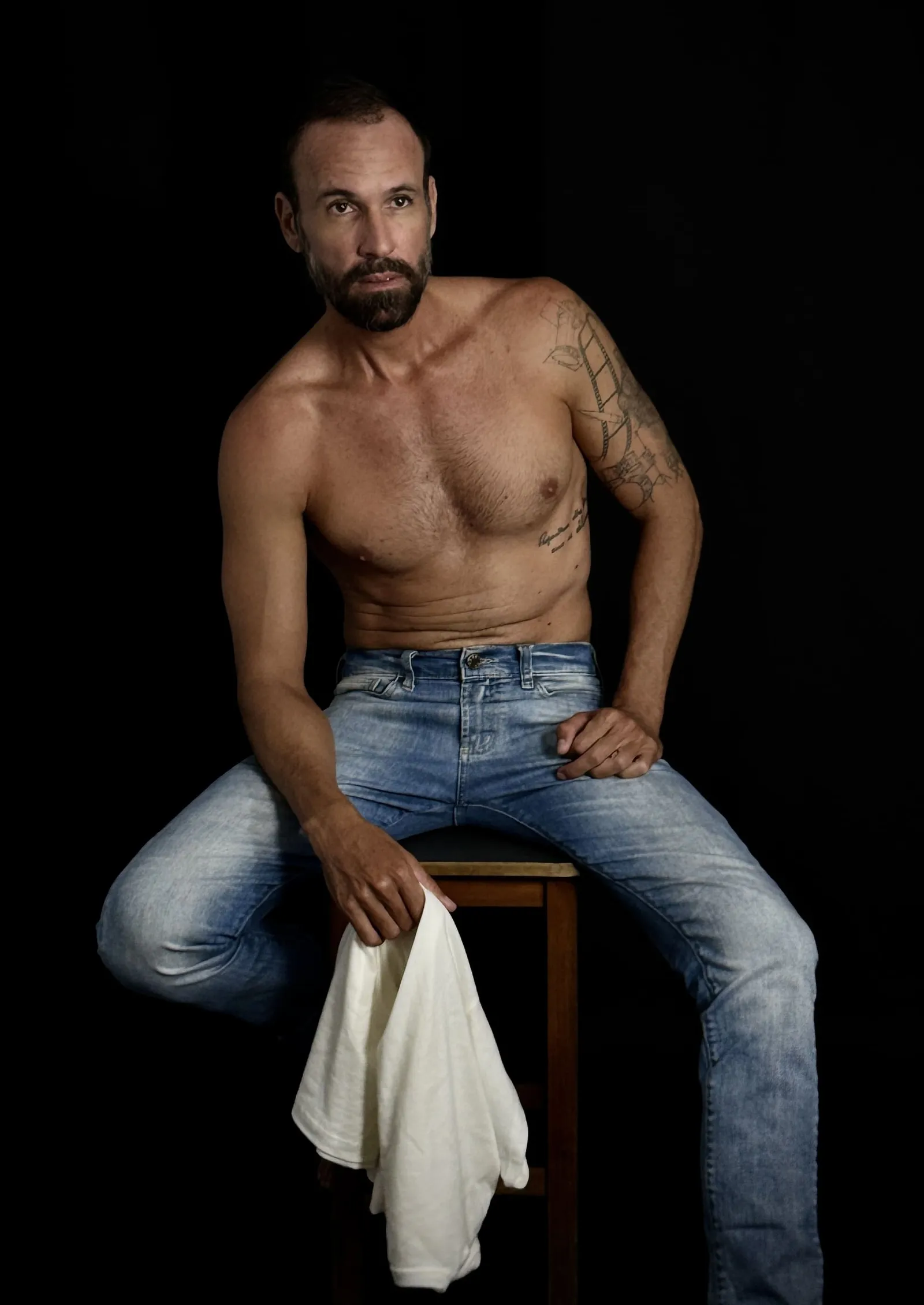 Vini Cavalieri sem camisa, de jeans, sentado num banco em fundo preto