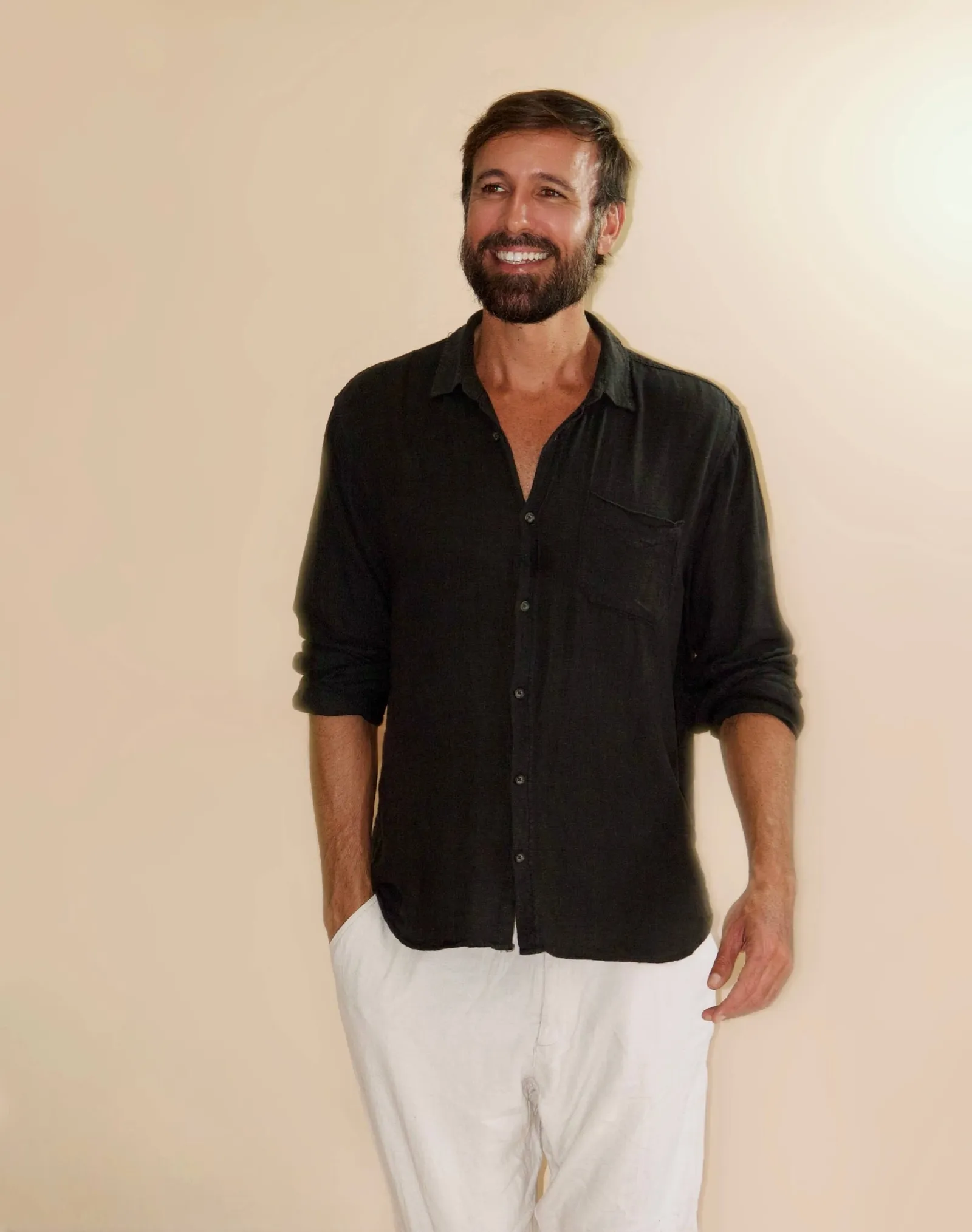 Vini Cavalieri sorrindo, de camisa preta e calça branca, em fundo bege