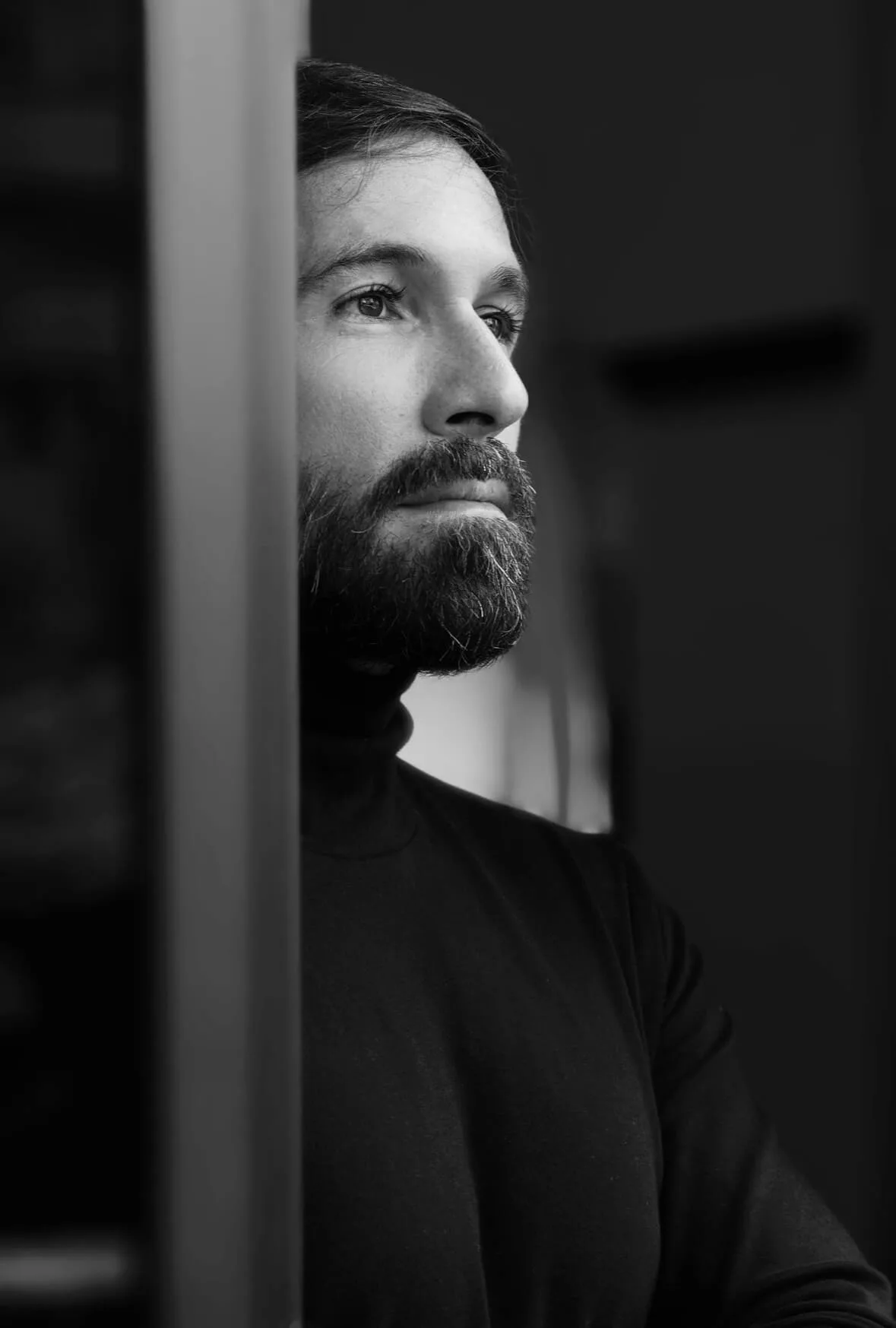 Retrato em preto e branco de Vini Cavalieri, de perfil junto a uma porta