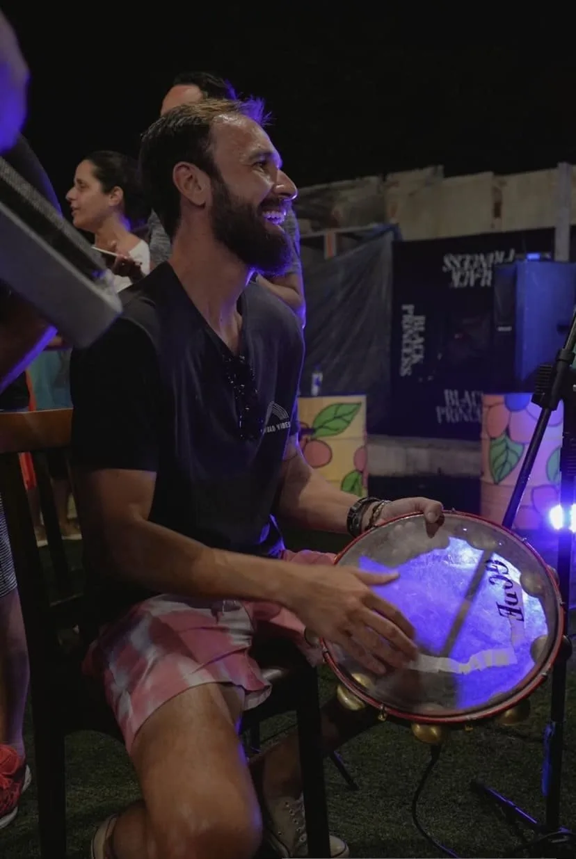 Vini Cavalieri sorrindo, tocando pandeiro numa roda de samba