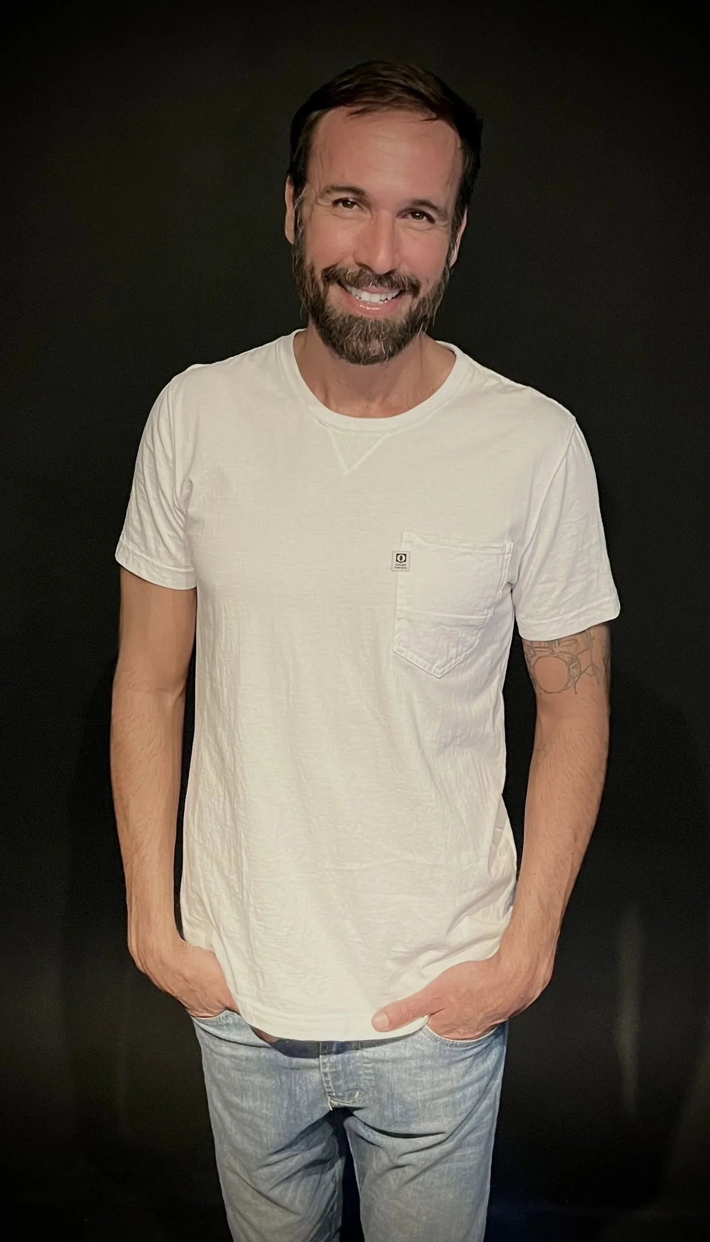 Vini Cavalieri sorrindo, de camiseta branca e jeans, em fundo preto