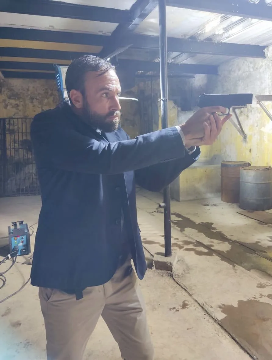 Vini Cavalieri apontando uma pistola, numa cena de ação num galpão