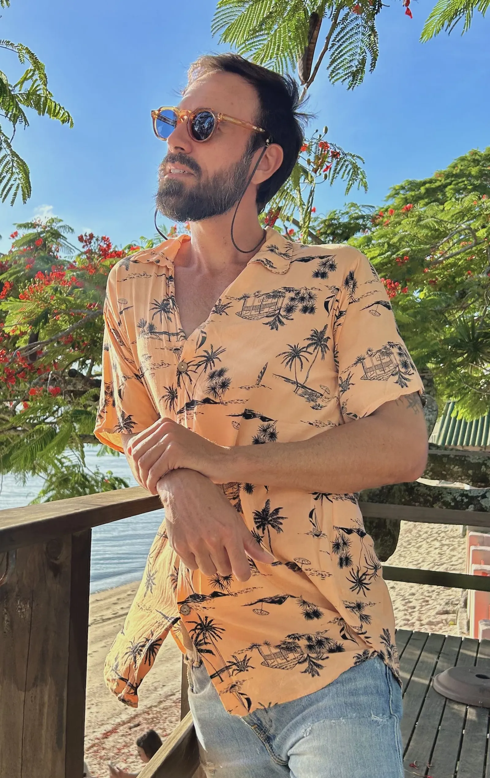 Vini Cavalieri de camisa estampada e óculos escuros, numa varanda com árvores floridas