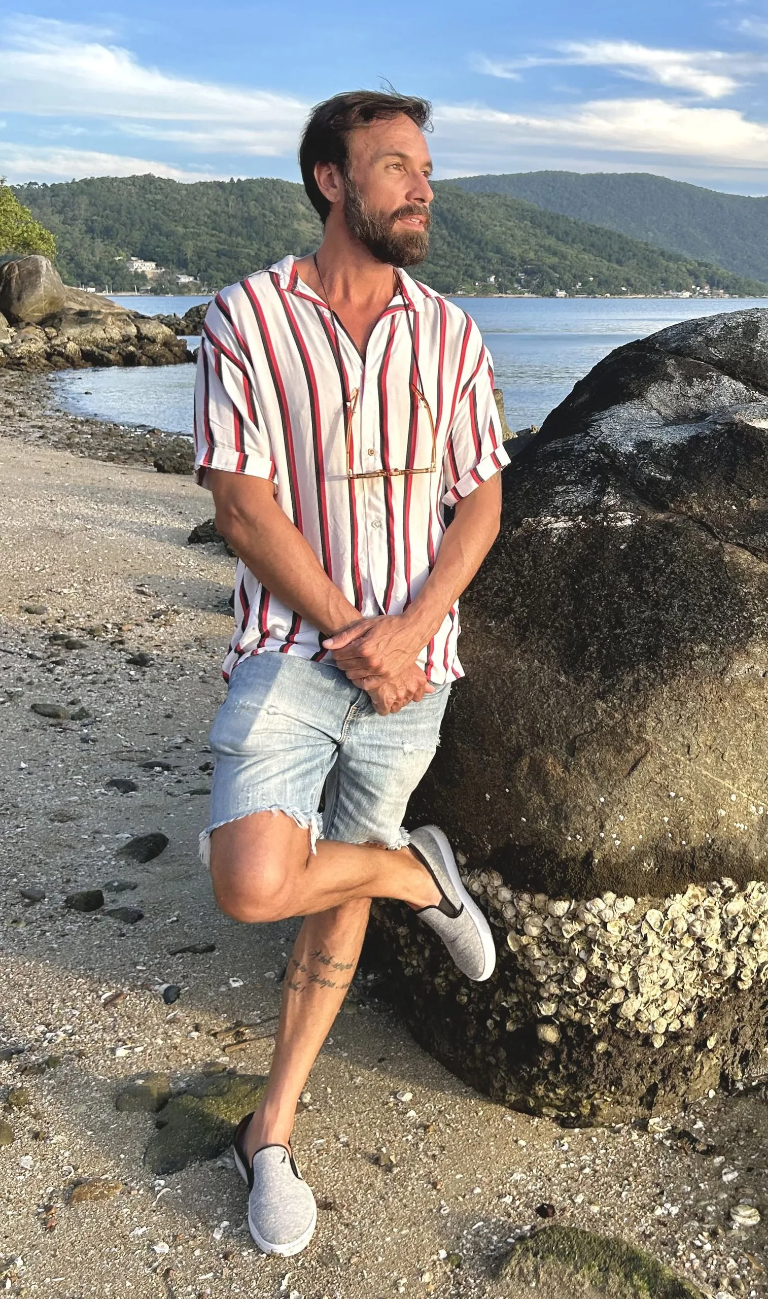 Vini Cavalieri de camisa listrada e bermuda, encostado a uma pedra na praia